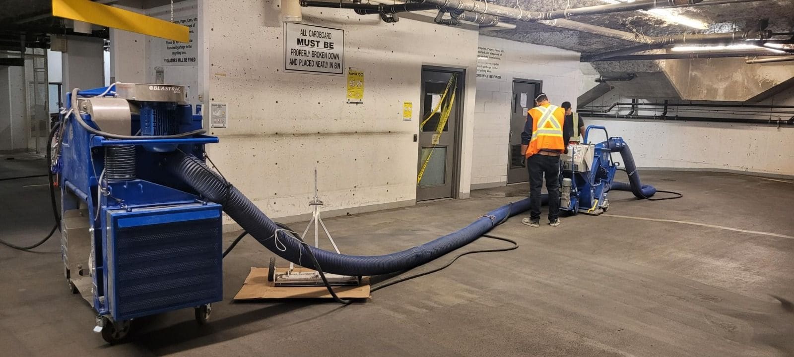 Two workers in hi-vis vests shot-blasting an underground parkade deck, a blue dust collector connected to the machine by hose